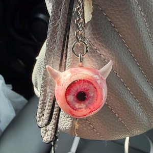 Puede incluir: Un llavero novedoso con un dije de ojo rosa con cuernos de diablo. El dije tiene un iris rojo y una pupila negra, con una cadena y un llavero plateados. El llavero está colgado de un bolso de cuero gris.