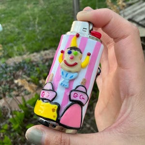 Puede incluir: Un encendedor decorativo con un diseño caprichoso. El encendedor presenta una figura de payaso con sombrero y brazos amarillos, una sonrisa roja y ojos azules. Rayas rosas y blancas adornan el cuerpo del encendedor, con dos diseños de pastel rosa y amarillo en la parte inferior.