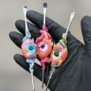 Peut inclure: Trois outils de sculpture en métal dotés de manches personnalisés en forme d'yeux réalistes avec des iris colorés en bleu, rose et vert. Chaque manche est orné de protubérances sculptées en forme de tentacules et de détails veineux autour de l'œil.