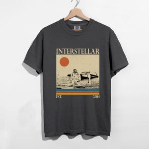 May include: Dark grey t-shirt with a retro graphic. The design features the word "INTERSTELLAR" above an astronaut in water with a spacecraft. Includes a sun and the text "EST. 2014".