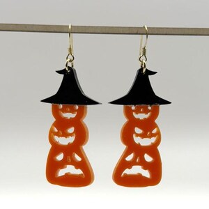 Op de afbeelding: Een paar Halloween-oorbellen. Elke oorbel heeft een gestapeld pompoenontwerp in oranje, bekroond met een zwarte heksenhoed. De pompoenen hebben uitgesneden gezichten. De oorbellen hangen aan goudkleurige haken.