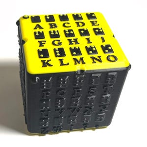 May include: A black and yellow cube with raised letters and Braille dots. The top is yellow with black letters A through N and Braille dots. The sides are black with raised letters and Braille. The cube is on a white background.