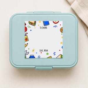 Peut inclure: Une boîte à lunch rectangulaire bleu clair avec un autocollant blanc carré sur le couvercle. L'autocollant présente une bordure de fournitures scolaires et d'aliments en dessin animé, avec le texte TO EMMA en haut et LOVE, MOM en bas.