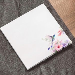 May include: A stack of white notepads with a watercolor hummingbird and floral design. The hummingbird has blue, green, and purple feathers, with pink, purple, and green flowers. The notepad is on a gray surface.