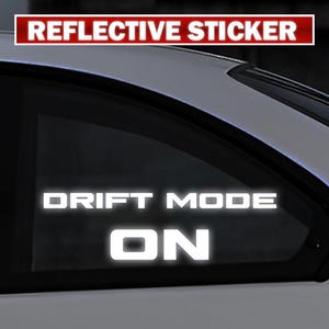 Puede incluir: Una pegatina reflectante blanca aplicada en la ventanilla de un coche con el texto DRIFT MODE ON en una fuente sans-serif en negrita. La pegatina está colocada en la ventanilla lateral trasera de un vehículo blanco con letras blancas.