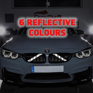 May include: A gray car with white reflective stripes on the front grill. The text "6 REFLECTIVE COLOURS" is displayed in red. The car has bright headlights and a license plate.
