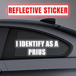 May include: A reflective sticker with the text "I IDENTIFY AS A PRIUS" in white, displayed on a car window. The sticker is above the text "REFLECTIVE STICKER" in a red rectangle.