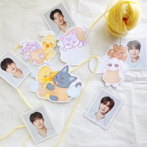 May include: A collection of stickers and photo cards featuring cartoon animals and portraits of people. The stickers include a cat, duck, and bear. The photo cards are rectangular and have a white border. A ball of yellow yarn is in the background.