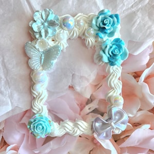 Pode incluir: Uma moldura decorativa branca com um design caprichoso. Apresenta flores azul claro, uma borboleta e contas iridescentes. A moldura tem uma borda ondulada e texturizada e um laço perolado. A moldura é cercada por tecido rosa e branco.