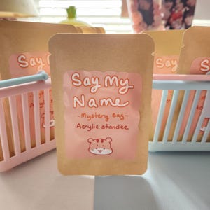 Puede incluir: Varias bolsas de papel marrón con etiquetas rosas que dicen "Say My Name - Mystery Bag - Acrylic standee". Las bolsas están en pequeñas cestas rosas y azules. Una linda ilustración de tigre está en la etiqueta.