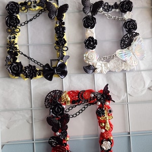 Pode incluir: Três molduras decorativas com enfeites de borboletas e rosas. Uma moldura é amarela com detalhes em preto, outra é branca com detalhes em preto e iridescentes, e a terceira é vermelha com detalhes em preto e dourado.