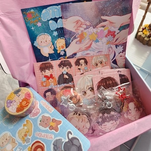 Puede incluir: Una caja de regalo rosa llena de varios artículos de papelería. Incluye pegatinas, cinta washi, postales y colgantes acrílicos con personajes de dibujos animados e ilustraciones de estilo anime. La caja está forrada con papel de seda rosa.