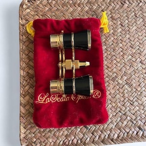May include: A pair of black and gold opera glasses resting on a red velvet pouch. The pouch has gold embroidered text that reads "La Scala Opus". The glasses are in a folded position.