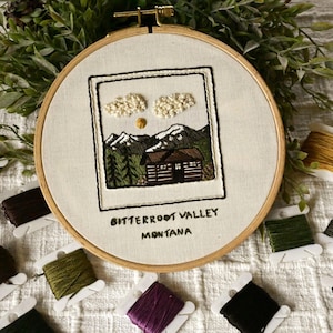 May include: Embroidered hoop art featuring a cabin scene with mountains, trees, and clouds. The text "BITTERROOT VALLEY MONTANA" is stitched below the image. The artwork is framed in a wooden hoop, with spools of thread around it.