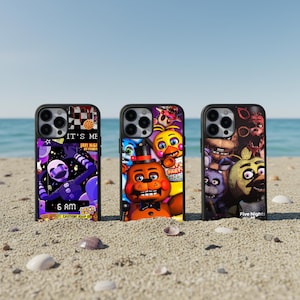 May include: Three black phone cases with colorful designs featuring characters and text from the video game series "Five Nights at Freddy's". The cases are on a sandy beach with the ocean in the background. The text on the cases includes "IT'S ME", "6 AM", and "Five Nights".