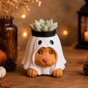 May include: A brown dog figurine wearing a white ghost costume with black eye holes. The top of the ghost costume features a black circular opening containing a small green succulent plant. The dog has a pink nose and visible front paws.
