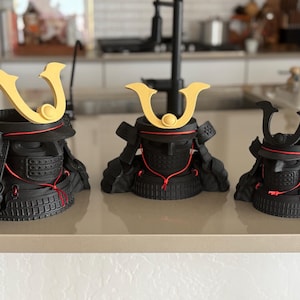 May include: Three black samurai helmet sculptures with gold crests and red accents. The helmets are displayed on a light-colored countertop. The helmets vary in size, showcasing a traditional Japanese design.
