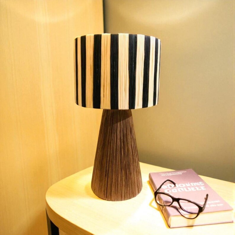 Op de afbeelding: Een decoratieve tafellamp met een cilindrische lampenkap met verticale zwarte en beige strepen. De lamp heeft een conische, bruine voet. Een boek en een bril liggen op de tafel.