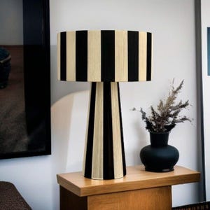 May include: A black and cream striped table lamp with a cylindrical lampshade and a conical base. The lamp is placed on a wooden table next to a black vase with dried plants. The lamp's design is modern and geometric.