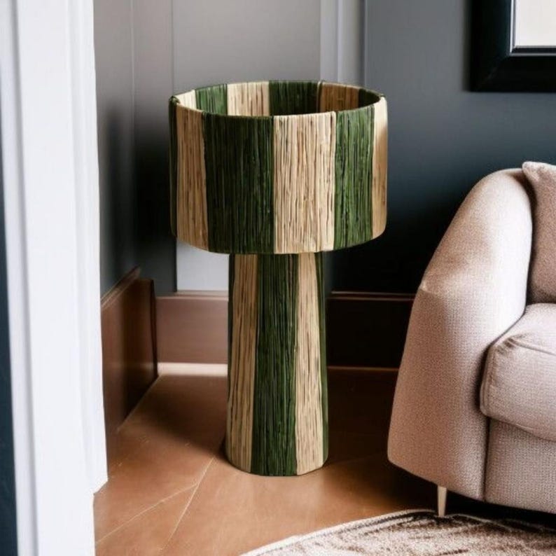 May include: A tall, decorative plant stand with a cylindrical base and a wide, open top. The stand is wrapped in vertical stripes of green and natural-colored woven material, creating a textured, striped pattern. The stand is approximately 30 inches tall.