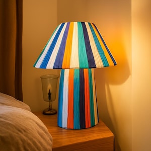 Könnte beinhalten: Eine farbenfrohe Tischlampe mit gestreiftem Lampenschirm und Sockel. Die Lampe hat vertikale Streifen in Blau, Orange, Grün und Weiß. Die Lampe steht auf einem Nachttisch aus Holz neben einem Glaskerzenhalter.