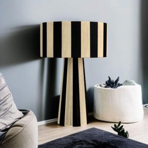 May include: A tall, modern floor lamp with a cylindrical lampshade and a conical base. The lampshade and base feature vertical black and beige stripes. The lamp is placed in a room with a gray wall, a dark rug, and a white cylindrical planter.