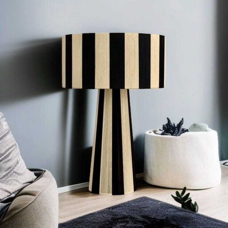 May include: A tall, modern floor lamp with a cylindrical lampshade and a conical base. The lampshade and base feature vertical black and beige stripes. The lamp is placed in a room with a gray wall, a dark rug, and a white cylindrical planter.