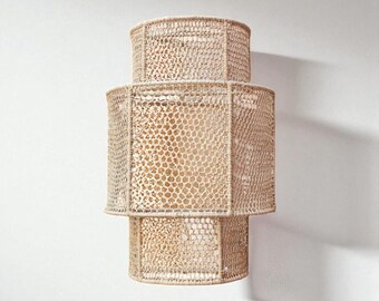 Op de afbeelding: Een beige, geweven wandlamp met een gelaagd ontwerp. De lamp heeft een zeshoekige vorm met een textuur, open weefpatroon. De lamp is bevestigd aan een witte muur en werpt een schaduw.