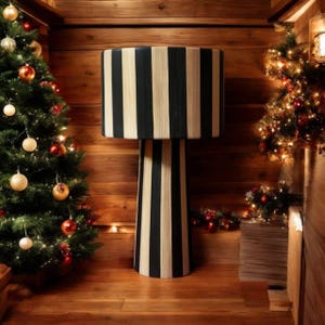 Op de afbeelding: Een hoge, zwart-beige gestreepte lamp staat in een gezellige, met hout beklede kamer, geflankeerd door kerstbomen versierd met ornamenten en lichtjes. De cilindrische kap en voet van de lamp hebben verticale strepen.