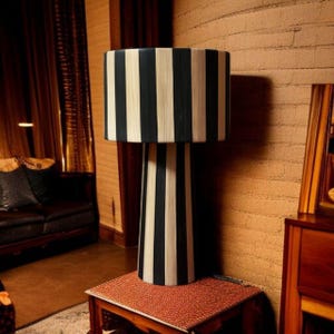Op de afbeelding: Een zwart-wit gestreepte tafellamp met een cilindrische kap en een conische voet. De lamp staat op een kleine, vierkante tafel met een rood patroon. De kamer heeft een warme, uitnodigende sfeer.