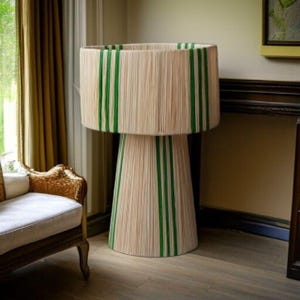 Puede incluir: Una l&aacute;mpara de pie grande, beige, con rayas verdes verticales. La l&aacute;mpara tiene una pantalla cil&iacute;ndrica y una base c&oacute;nica, ambas adornadas con rayas verdes. La l&aacute;mpara est&aacute; hecha de un material tejido, creando una apariencia texturizada. Es una l&aacute;mpara decorativa.