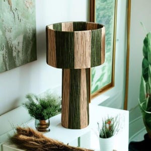 May include: A decorative table lamp with a cylindrical base and shade. The lamp features vertical stripes of olive green and beige. The lamp is placed on a white surface, with small plants in the foreground and a painting in the background.
