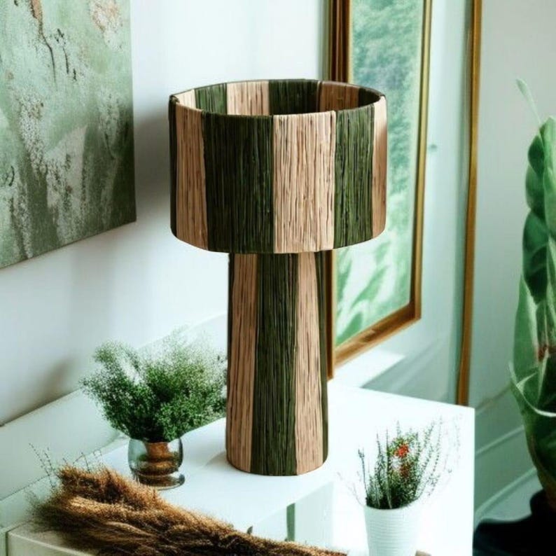 May include: A decorative table lamp with a cylindrical base and shade. The lamp features vertical stripes of olive green and beige. The lamp is placed on a white surface, with small plants in the foreground and a painting in the background.