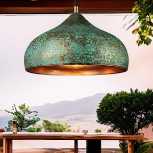 Pode incluir: Uma luminária pendente turquesa e dourada com uma superfície texturizada. A luz tem um interior cor de cobre e é suspensa por um cabo preto com um detalhe dourado. A luz tem formato de cúpula e está pendurada sobre uma mesa de madeira.