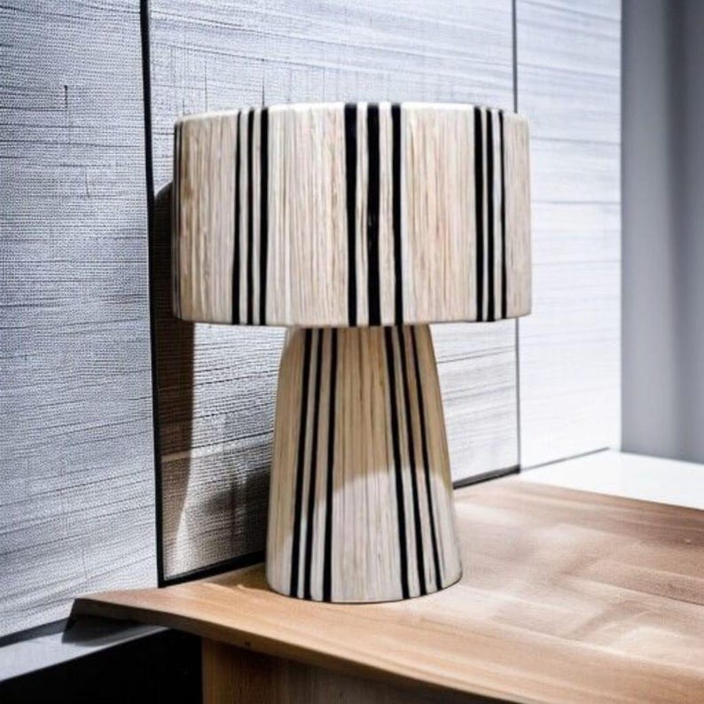 Puede incluir: Una l&aacute;mpara de mesa decorativa con una pantalla cil&iacute;ndrica y una base c&oacute;nica. La l&aacute;mpara presenta un color beige natural con rayas negras verticales. La l&aacute;mpara est&aacute; colocada sobre una mesa de madera.