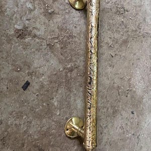 Könnte beinhalten: Ein goldfarbener Türgriff mit aufwendigen Blumen- und Blattmustern. Der Griff ist vertikal ausgerichtet, mit dekorativen Zierknöpfen an jedem Ende und kreisförmigen Montageplatten.