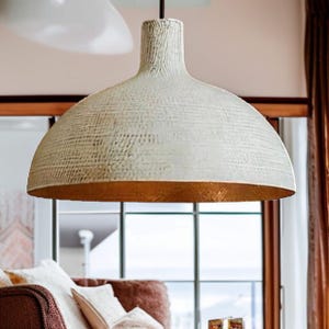 May include: Cream-colored, textured pendant light with a wide, dome-shaped shade. The interior of the shade has a gold finish. The light hangs from a black cord and is suspended above a living room setting.