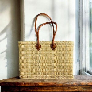 K&ouml;nnte beinhalten: Eine rechteckige Stroh-Tragetasche mit braunen Ledergriffen. Die Tasche ist hellbeige und steht auf einer Holzoberfl&auml;che. Die Griffe sind mit herzf&ouml;rmigen Lederaufn&auml;hern befestigt.