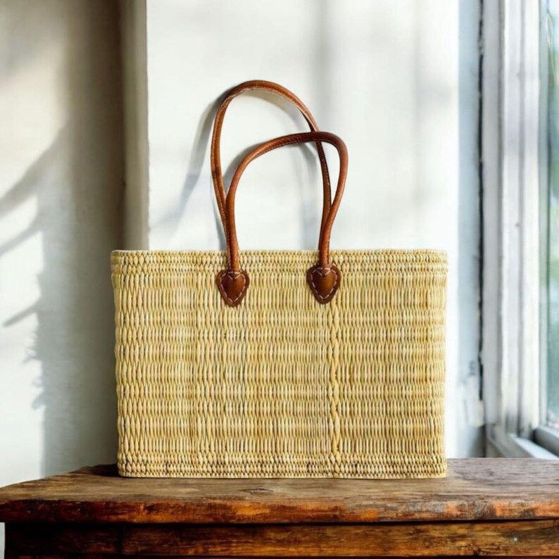 K&ouml;nnte beinhalten: Eine rechteckige Stroh-Tragetasche mit braunen Ledergriffen. Die Tasche ist hellbeige und steht auf einer Holzoberfl&auml;che. Die Griffe sind mit herzf&ouml;rmigen Lederaufn&auml;hern befestigt.