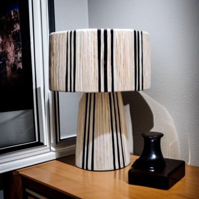Puede incluir: L&aacute;mpara de mesa decorativa con pantalla y base cil&iacute;ndricas, ambas adornadas con rayas negras verticales sobre un fondo crema. Un peque&ntilde;o objeto negro con base cuadrada se encuentra sobre la mesa de madera junto a la l&aacute;mpara.