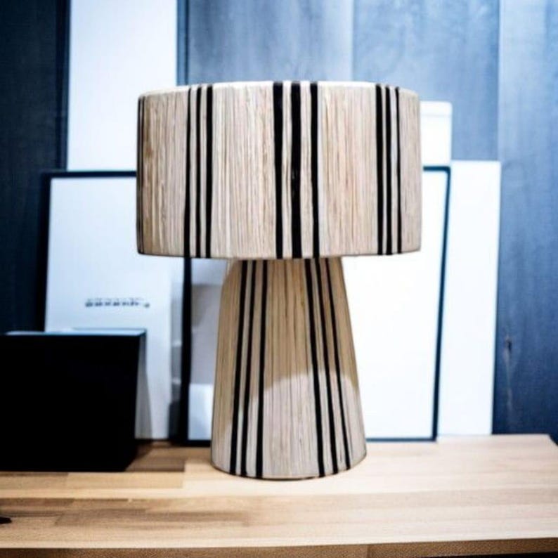 Puede incluir: Una l&aacute;mpara de mesa decorativa con una pantalla cil&iacute;ndrica y una base c&oacute;nica. La l&aacute;mpara presenta una textura tejida beige natural con rayas negras verticales. La l&aacute;mpara est&aacute; colocada sobre una superficie de madera.