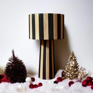 Könnte beinhalten: Eine dekorative Lampe mit zylindrischem Lampenschirm und konischem Sockel. Die Lampe hat vertikale schwarze und natürliche Holzstreifen. Die Lampe ist von kleinen Weihnachtsbäumen, roten Kugeln und weißem Kunstfell umgeben.