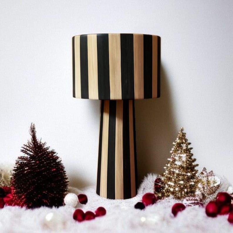 Op de afbeelding: Een decoratieve lamp met een cilindrische lampenkap en een conische voet. De lamp heeft verticale zwarte en natuurlijke houten strepen. De lamp is omgeven door kleine kerstbomen, rode ballen en witte imitatiebont.