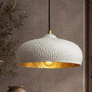 May include: A dome-shaped pendant light fitting featuring a white exterior with a textured, hammered finish. The interior of the shade has a metallic gold finish, and the fitting is suspended by a black cord with a brass-coloured metal connector.