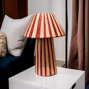 Könnte beinhalten: Eine dekorative Tischlampe mit einem rot-beige gestreiften Schirm und Sockel. Die Lampe ist zylindrisch und steht auf einer weißen Oberfläche. Das Design der Lampe ist eine Mischung aus modernem und Retro-Stil.