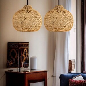 May include: Two woven rattan pendant lights suspended from the ceiling. The lamps have a round, bulbous shape with a woven pattern. The lights are suspended by black cords. The background includes a wooden table, artwork, and a blue sofa.