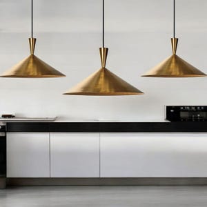 K&ouml;nnte beinhalten: Drei Pendelleuchten aus Messing mit konischen Schirmen h&auml;ngen &uuml;ber einer wei&szlig;en und schwarzen K&uuml;chenzeile. Die Leuchten haben ein schlankes, modernes Design mit einer schmalen, spitzen Oberseite und einer breiten, ausgestellten Basis. Die K&uuml;chenschr&auml;nke sind wei&szlig; mit schwarzen Arbeitsplatten.