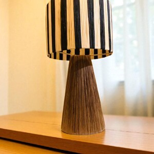 Op de afbeelding: Decoratieve tafellamp met een cilindrische lampenkap met verticale zwarte en beige strepen. De voet van de lamp heeft een conische vorm in bruin, wat een natuurlijk, gestructureerd uiterlijk cre&euml;ert. De lamp staat op een houten oppervlak.