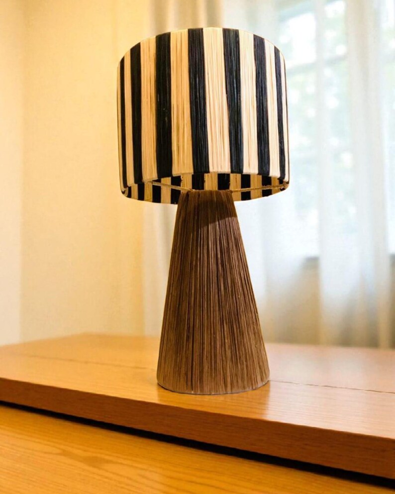 Op de afbeelding: Decoratieve tafellamp met een cilindrische lampenkap met verticale zwarte en beige strepen. De voet van de lamp heeft een conische vorm in bruin, wat een natuurlijk, gestructureerd uiterlijk cre&euml;ert. De lamp staat op een houten oppervlak.