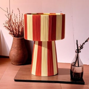 May include: A decorative table lamp with a cylindrical shade and base, featuring vertical stripes in red and cream. The lamp is made of yarn or thread, creating a textured surface. The lamp sits on a dark brown wooden base.
